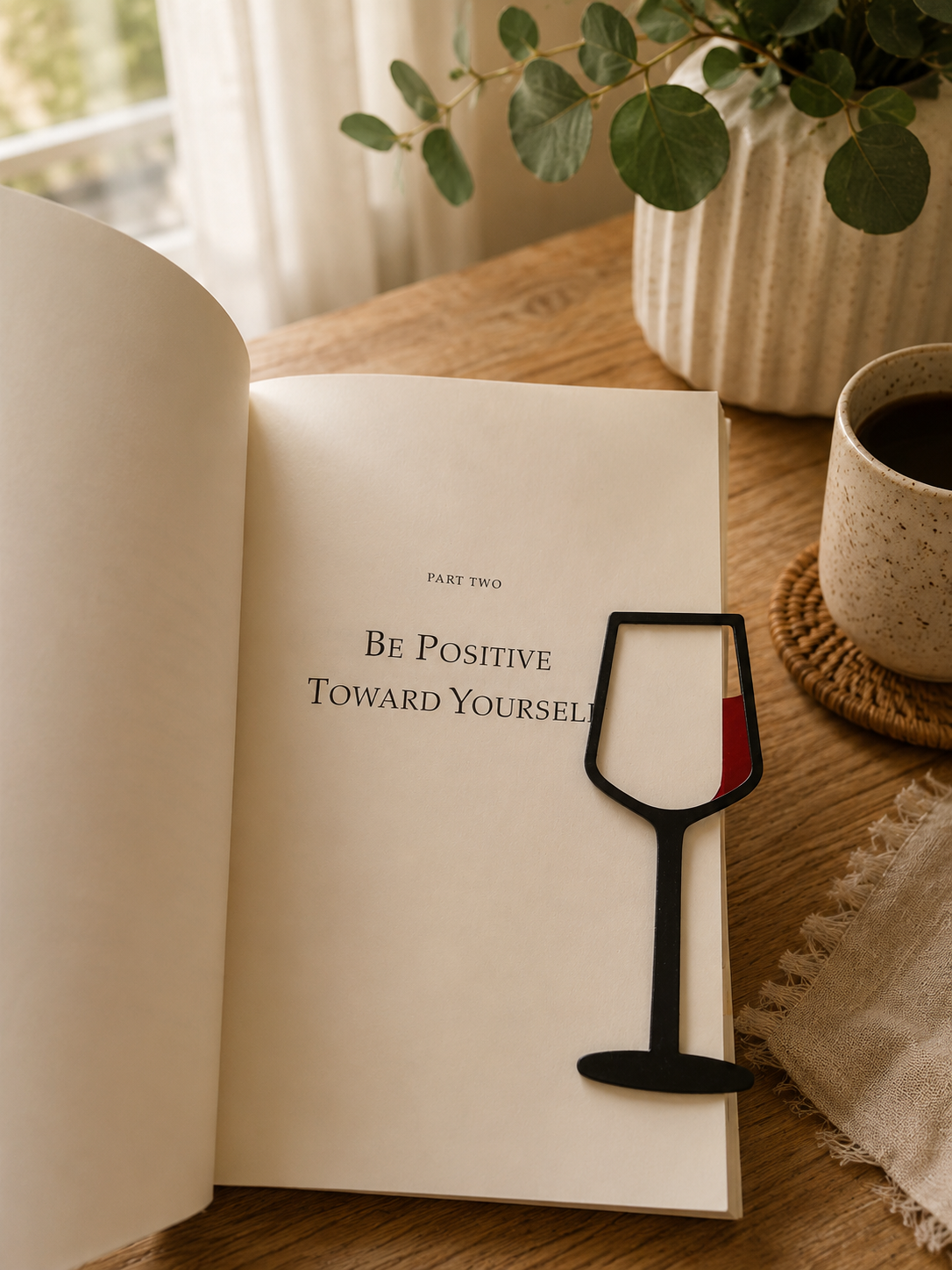 Black wine glass bookmark 3D printed, resting on an open book beside coffee and eucalyptus on a wooden table